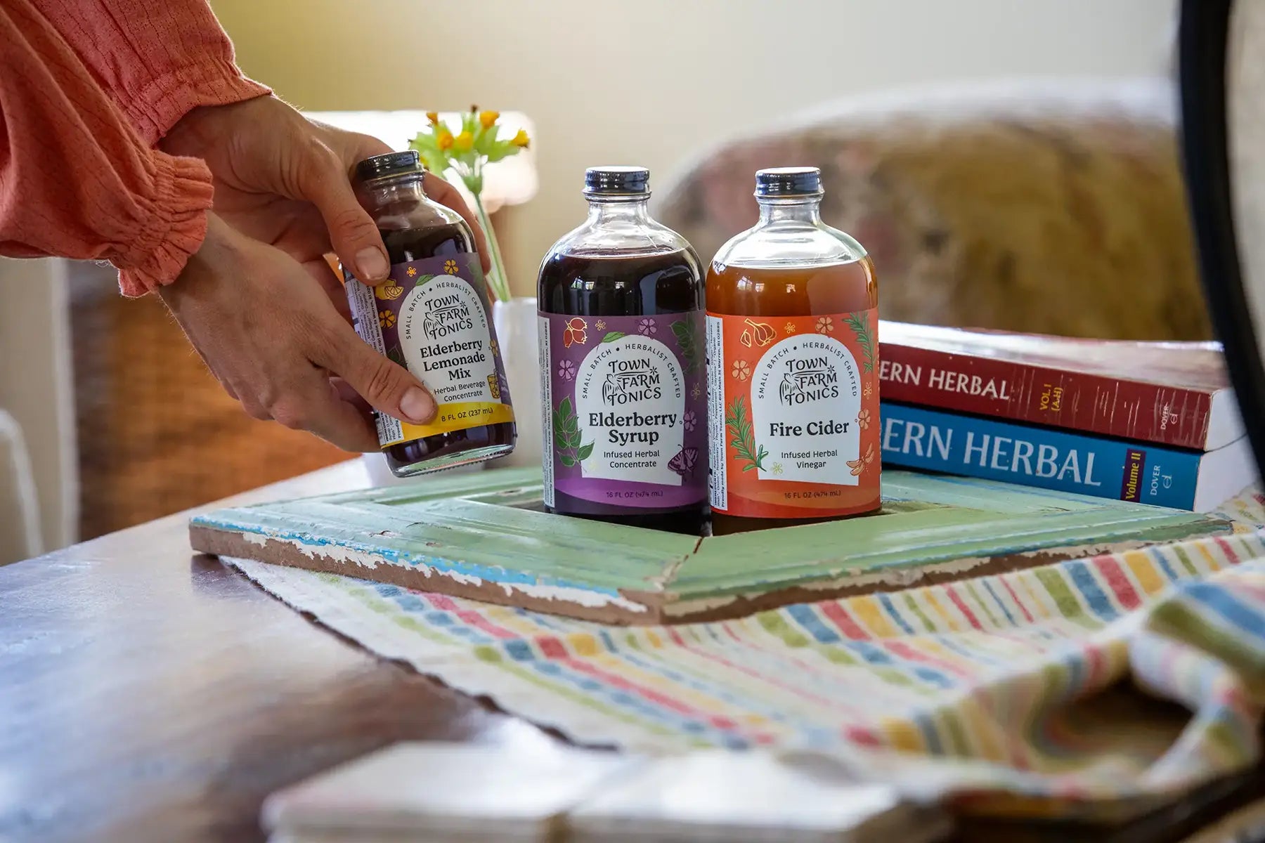 Bottles of herbal tonic products on a table with a colorful towel and books in the background.