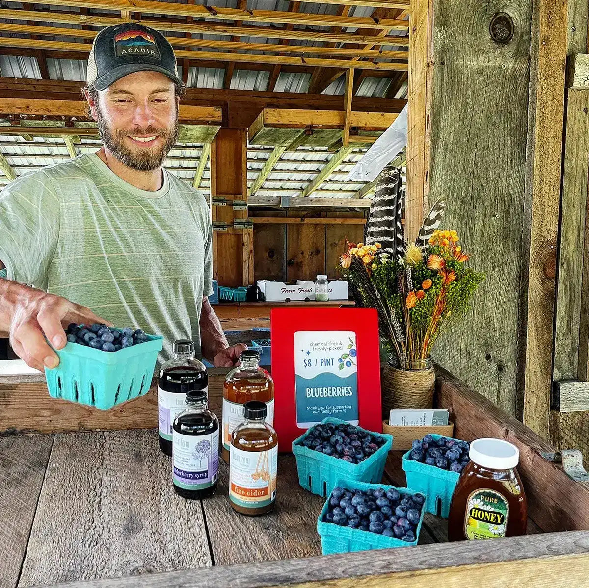 Owner holding a basket of blueberries with various products on a wooden counter.