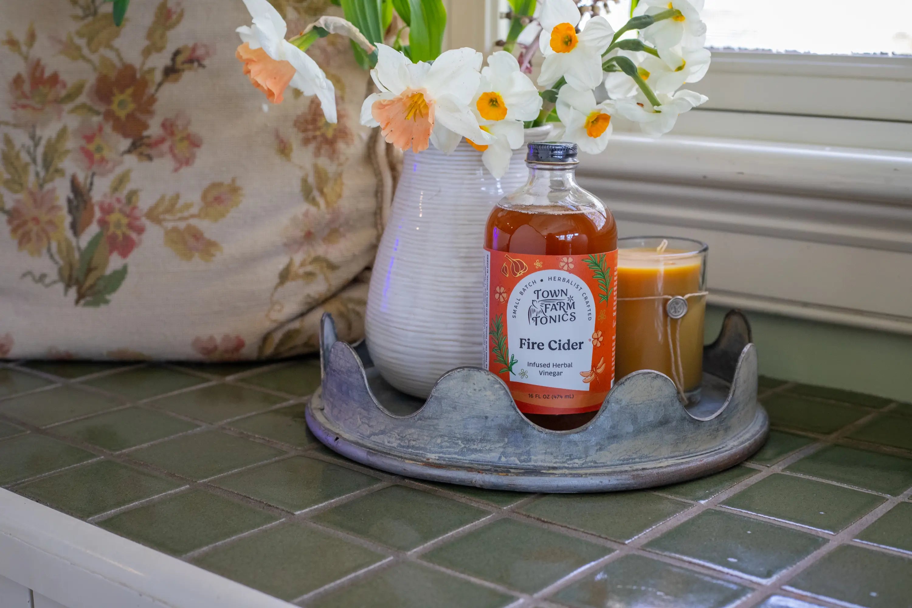 Bottle of Town Farm Fire Cider on a tray with flowers and a candle on a tiled surface.