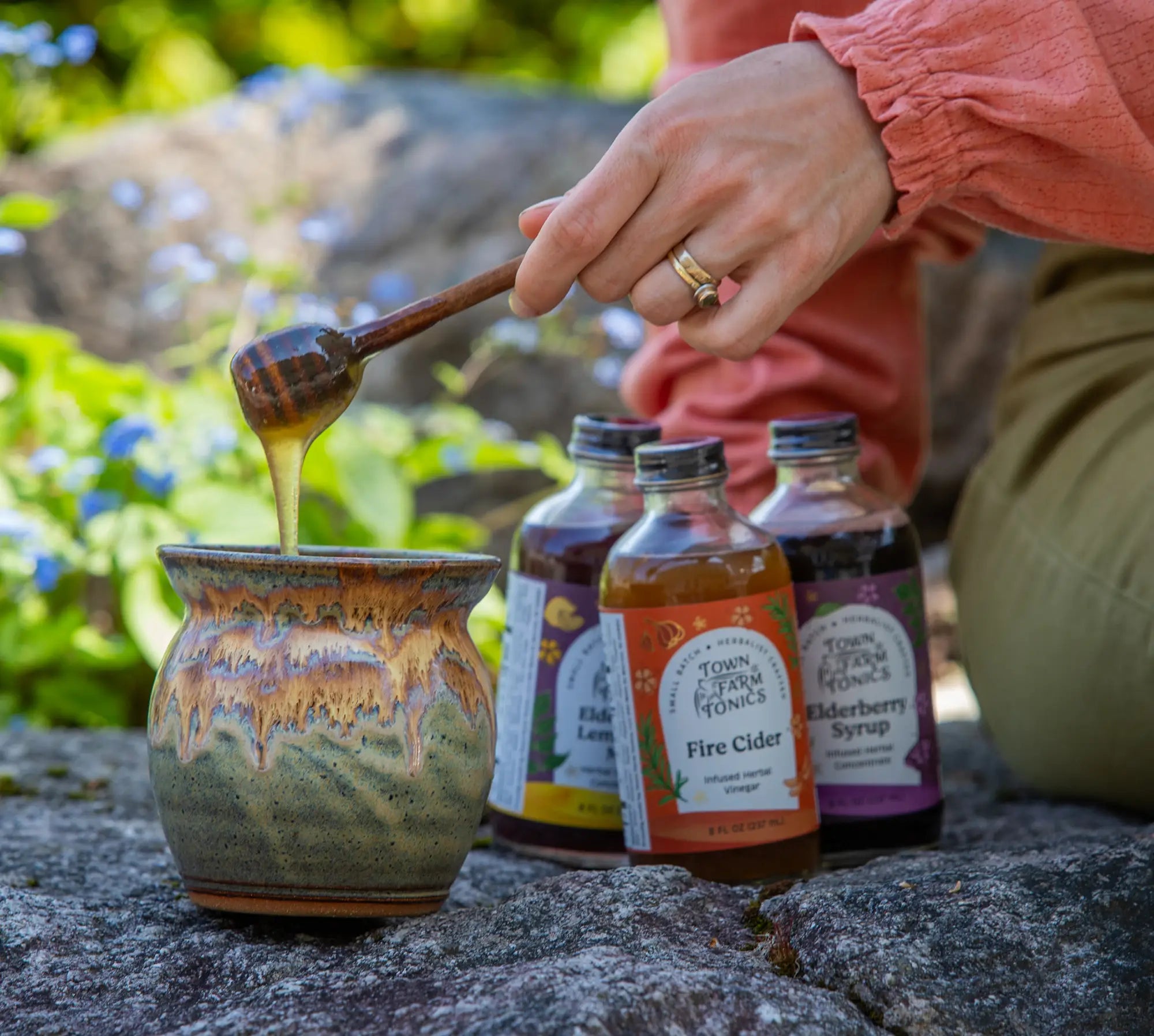 Person pouring honey from a wooden spoon into a ceramic jar with bottles of Town Farm Tonic in the background.