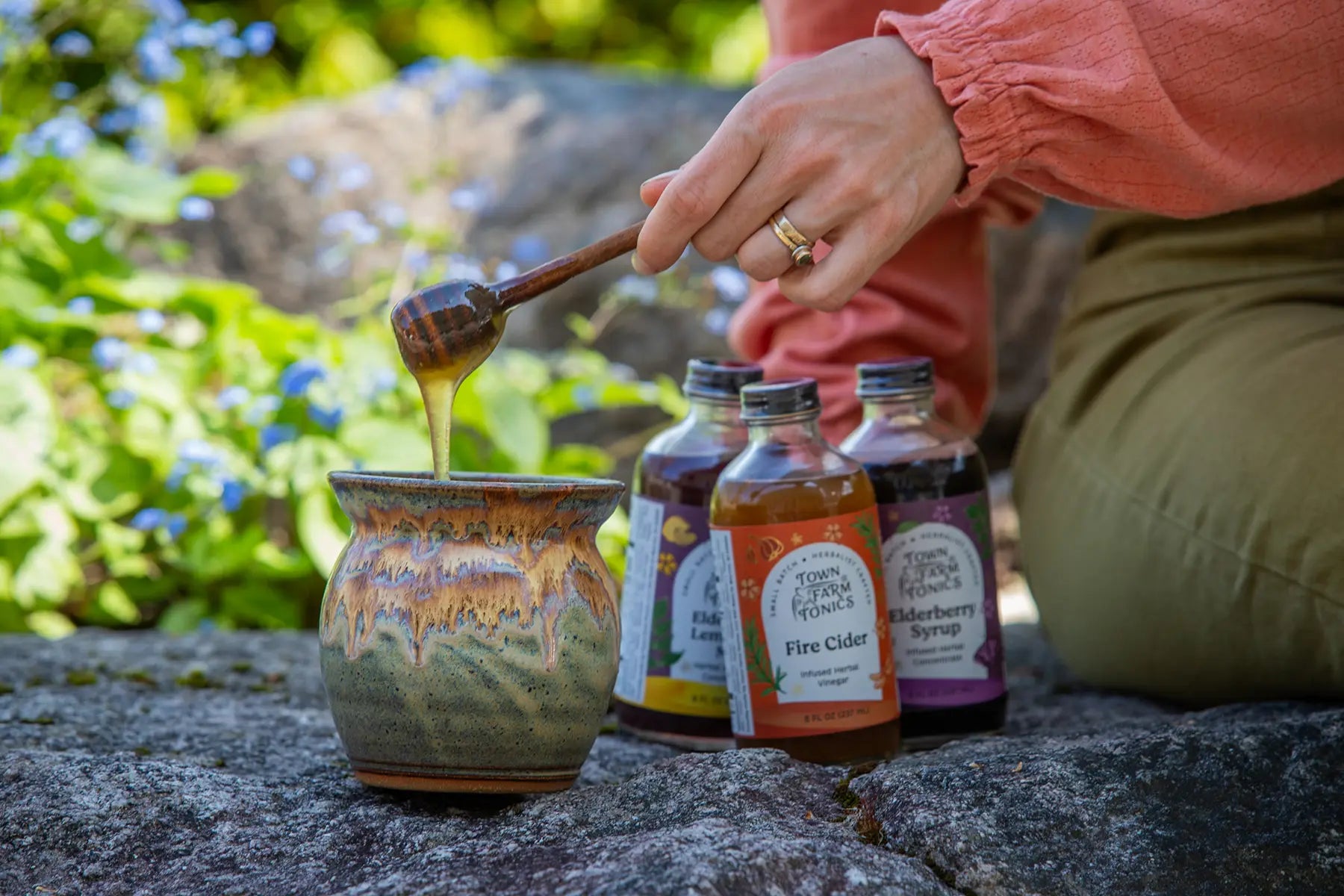 Person pouring honey into a jar with bottles of tonics in the background.