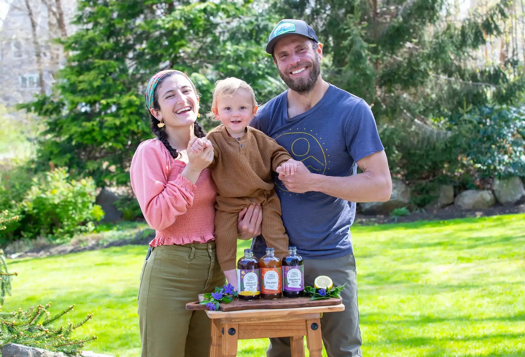 Owners and baby standing outdoors with a wooden table displaying tonic bottles